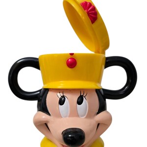 May include: A yellow Minnie Mouse-shaped cup with black handles and a lid. The cup features Minnie's face with a black nose, pink cheeks, and large eyes. The lid is open, revealing the cup's interior.