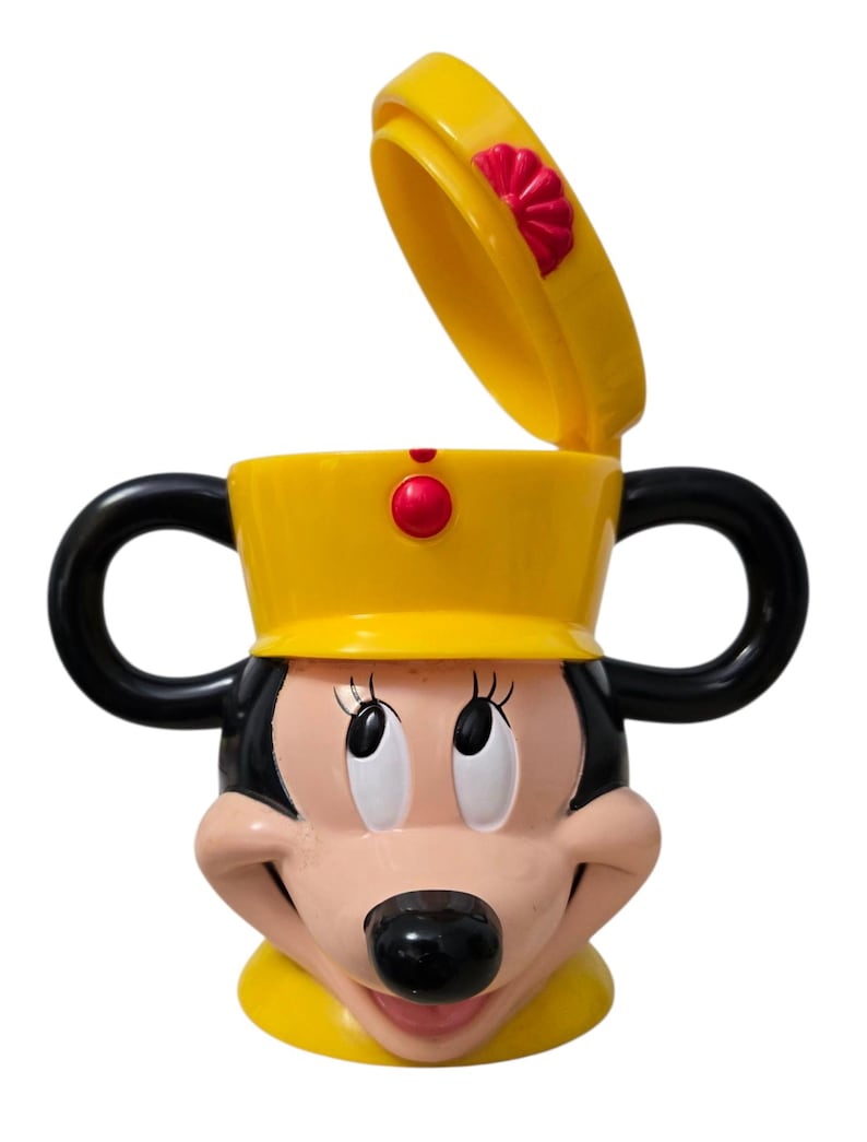 May include: A yellow Minnie Mouse-shaped cup with black handles and a lid. The cup features Minnie's face with a black nose, pink cheeks, and large eyes. The lid is open, revealing the cup's interior.