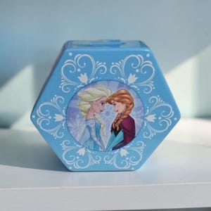 Puede incluir: Una hucha hexagonal de metal azul claro con un gráfico circular de dos personajes animados con cabello rubio y castaño. La caja está decorada con patrones de volutas florales blancas y un diseño de copo de nieve en el fondo de la imagen central.