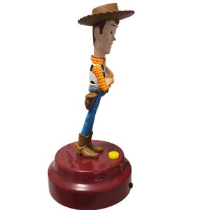 May include: A Woody bobblehead figurine. The figure is wearing a brown cowboy hat, a yellow plaid shirt, a white vest, blue pants, and brown boots. The base is a maroon color with a yellow button.