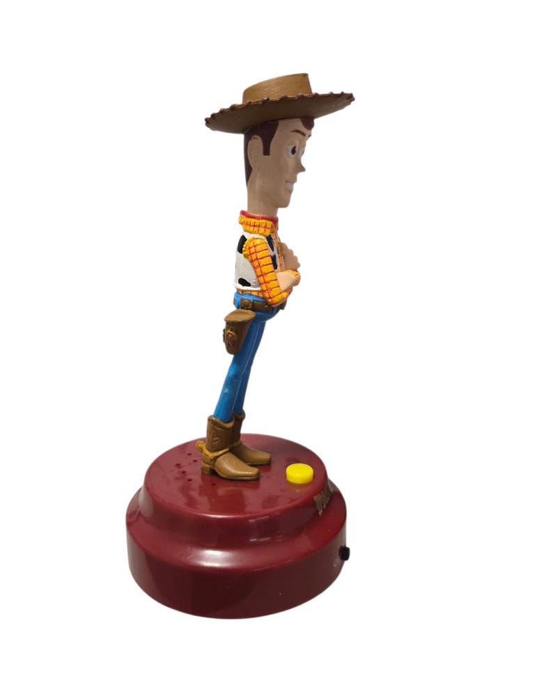 May include: A Woody bobblehead figurine. The figure is wearing a brown cowboy hat, a yellow plaid shirt, a white vest, blue pants, and brown boots. The base is a maroon color with a yellow button.