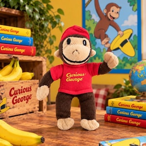 May include: A plush Curious George monkey toy wearing a red shirt and a red hat. The monkey has brown fur on its arms and legs with tan hands, feet, and face. The shirt features the text Curious George printed in yellow on the front chest area.