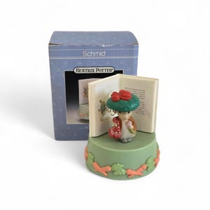 Puede incluir: Una figura de Beatrix Potter con una base verde y un fondo en forma de libro. La figura representa un conejo con un sombrero verde con un lazo rojo y sosteniendo un vestido rojo. La base está decorada con zanahorias y hojas. La caja dice "Schmid" y "Beatrix Potter."