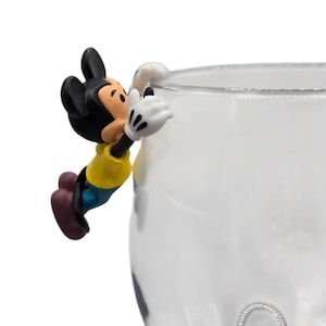 May include: A novelty glass with a decorative Mickey Mouse figure clinging to the rim. The figure is in a yellow shirt, blue shorts, and black ears. The glass is clear with a silver-colored, circular design on the side.