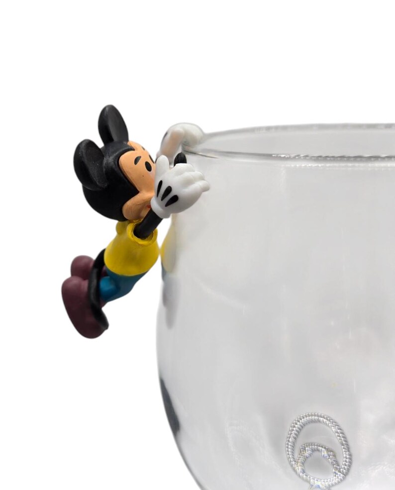May include: A novelty glass with a decorative Mickey Mouse figure clinging to the rim. The figure is in a yellow shirt, blue shorts, and black ears. The glass is clear with a silver-colored, circular design on the side.