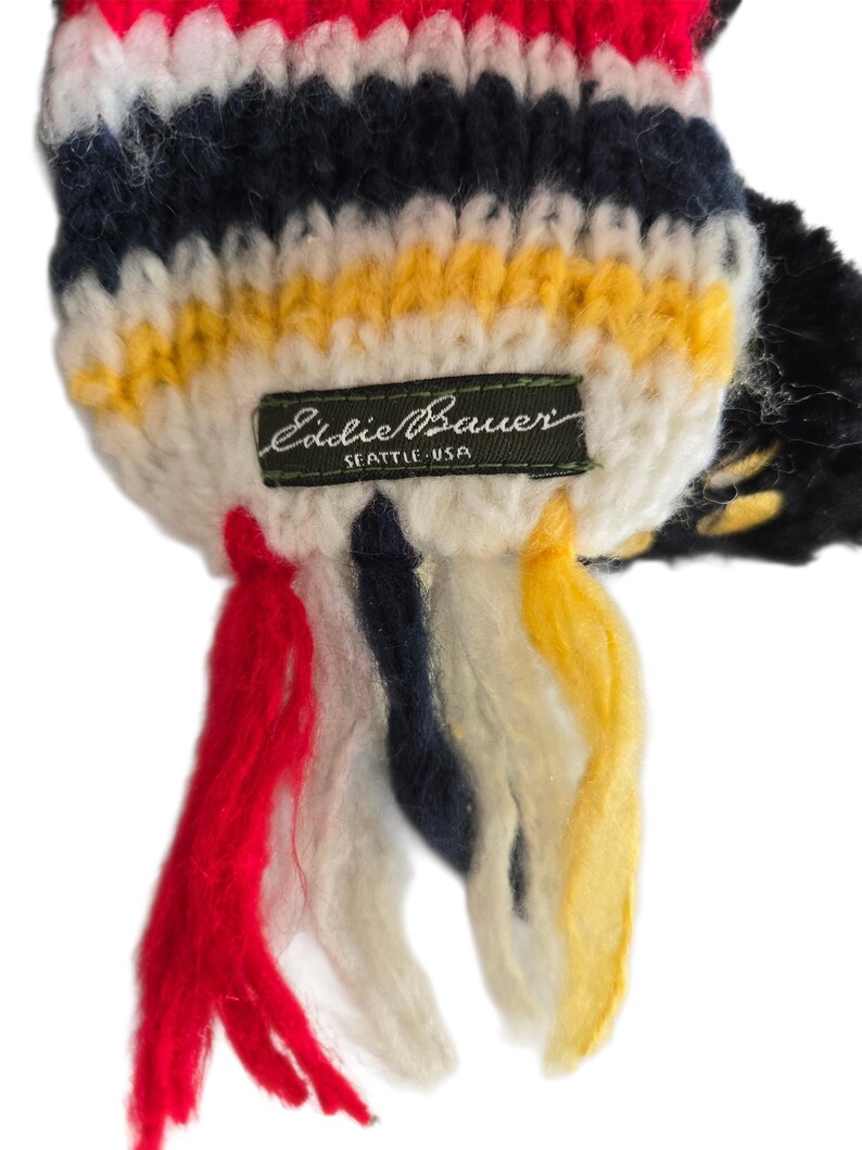 May include: Close-up of a colorful knit scarf with red, white, navy, and yellow stripes. The scarf has a tag that reads "Eddie Bauer SEATTLE USA" and features red, navy, and yellow tassels.