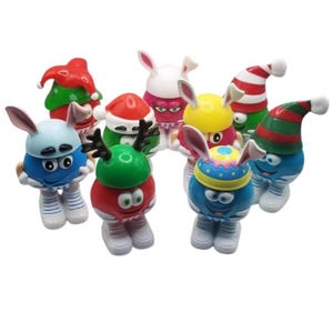 May include: A group of colourful, anthropomorphic candy characters, each with bunny ears and festive hats. The characters are in various colours, including red, blue, green, and yellow, with white shoes. Some wear Santa hats, others Easter bonnets, and some have reindeer antlers.