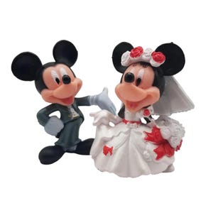 May include: Figurines of Mickey and Minnie Mouse in wedding attire. Mickey wears a dark green suit and Minnie a white wedding dress with red accents. Minnie has a veil and a floral headpiece.