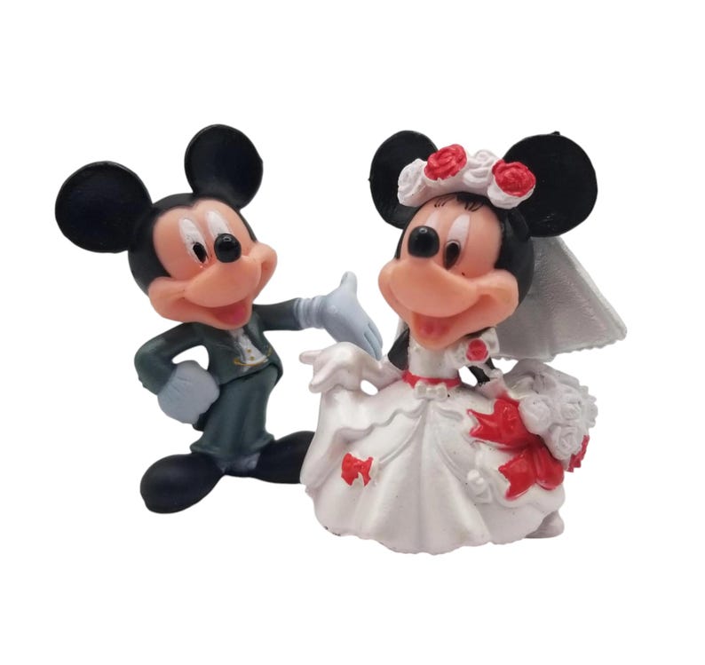 May include: Figurines of Mickey and Minnie Mouse in wedding attire. Mickey wears a dark green suit and Minnie a white wedding dress with red accents. Minnie has a veil and a floral headpiece.