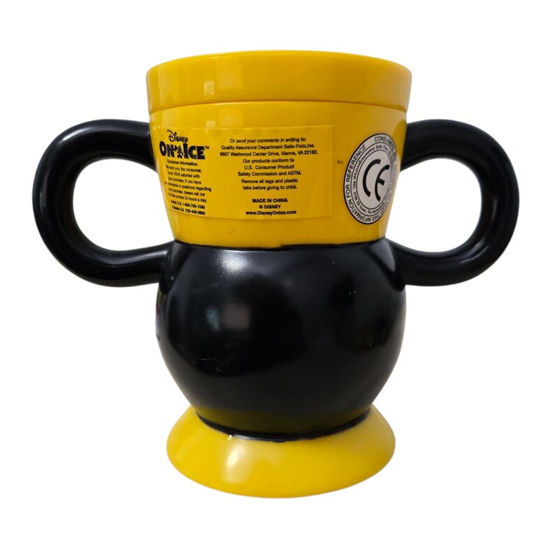 May include: A yellow and black Disney On Ice mug with two black handles. The mug has a rounded base and a cylindrical top. The mug has text on the side, including the Disney On Ice logo and other information.