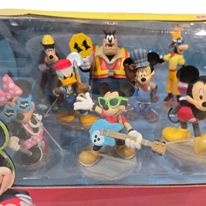 May include: A collection of Disney character figurines, including Mickey Mouse, Minnie Mouse, Donald Duck, Goofy, and others. The figures are in various poses and outfits, with vibrant colors and detailed designs. The set is packaged in a clear plastic container.