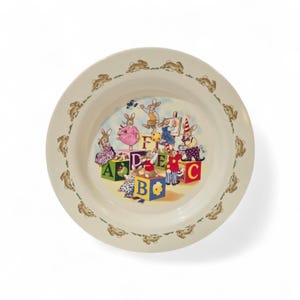 May include: A cream ceramic bowl with a rim decorated with brown rabbit silhouettes. The bowl's interior features a colourful illustration of rabbits playing with alphabet blocks. The blocks spell out letters A, B, C, D, E, and F.