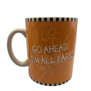 以下が含まれることがあります： 白いハンドルと「GO AHEAD. I'M ALL EARS」の白い文字が入ったオレンジ色のセラミックマグ。上部と下部の縁には白と黒のチェック柄があります。マグカップの内側は薄いベージュ色です。
