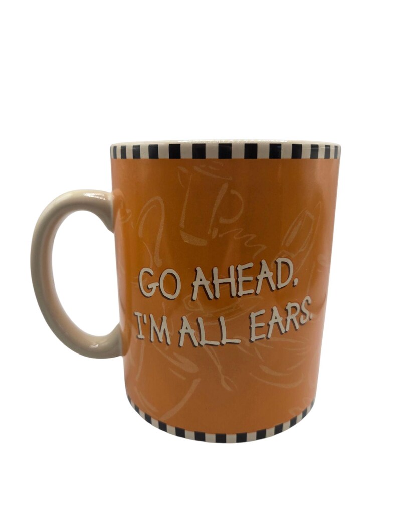 以下が含まれることがあります： 白いハンドルと「GO AHEAD. I'M ALL EARS」の白い文字が入ったオレンジ色のセラミックマグ。上部と下部の縁には白と黒のチェック柄があります。マグカップの内側は薄いベージュ色です。