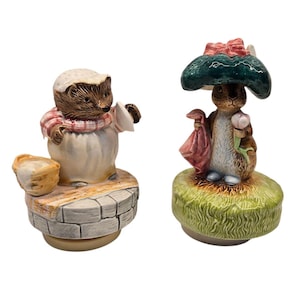 Puede incluir: Dos figuras de cerámica. Una es un erizo con gorro y delantal de chef, de pie sobre un pozo de piedra. La otra es un conejo bajo un paraguas de seta, sosteniendo una bolsa y una flor. Ambas sobre fondo blanco.