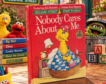 May include: A red hardcover children's book titled Nobody Cares About Me featuring Big Bird in a blue and white checkered blanket sitting in a basket. The cover shows a brick wall background, a lamp, an alarm clock, and a tissue box.