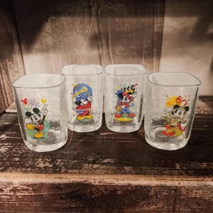 May include: Four clear glass tumblers with rounded square rims, each featuring a colourful graphic of Mickey Mouse in different outfits, including a magician, a director, and a festive celebration theme with fireworks and confetti patterns.