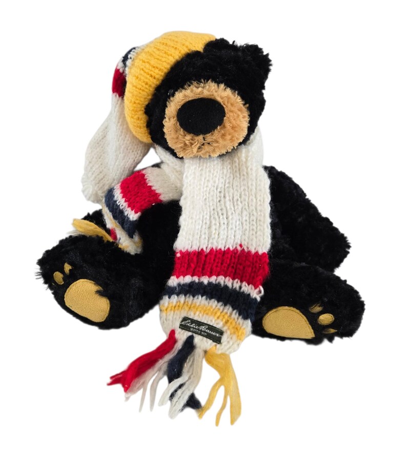 May include: A black teddy bear wearing a knit hat and scarf. The hat is yellow, red, and white. The scarf is white with red, navy, and yellow stripes. The bear has tan paws and a tan muzzle.