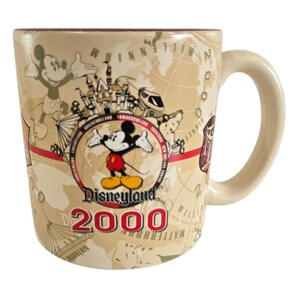Pode incluir: Caneca de cer&acirc;mica bege Disneyland 2000 com Mickey Mouse, um design de mapa e atra&ccedil;&otilde;es da Disneyland. A caneca tem uma al&ccedil;a curva e uma borda marrom. O texto "Disneyland 2000" &eacute; exibido em vermelho.