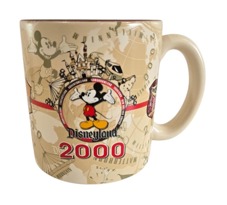 Pode incluir: Caneca de cer&acirc;mica bege Disneyland 2000 com Mickey Mouse, um design de mapa e atra&ccedil;&otilde;es da Disneyland. A caneca tem uma al&ccedil;a curva e uma borda marrom. O texto "Disneyland 2000" &eacute; exibido em vermelho.