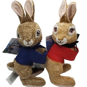 May include: Two plush Peter Rabbit toys. The rabbits have brown and tan fur, long ears, and embroidered facial features. One wears a blue shirt, the other a red shirt. Each has a tag attached.