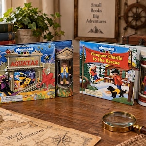以下が含まれることがあります： Playmobil Booksのセット「Adventure in Aquatax」と「Chopper Charlie to the Rescue」の2点。それぞれ絵本と、小さなプラスチック製のフィギュアが入った透明な円筒形のケースがセットになっています。木製のテーブルの上に置かれています。