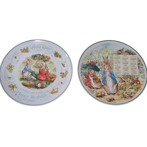 Puede incluir: Dos platos decorativos con ilustraciones de Peter Rabbit. Uno muestra conejos y el texto "Happy Birthday 2000". El otro tiene un calendario e ilustraciones de conejos. Ambos platos tienen un borde azul.