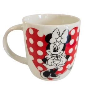 Op de afbeelding: Witte keramische mok met Minnie Mouse. De mok heeft een rood hartontwerp met witte stippen. Minnie Mouse is afgebeeld in zwart, wit en rood, met haar handen voor zich. De mok heeft een gebogen handvat.