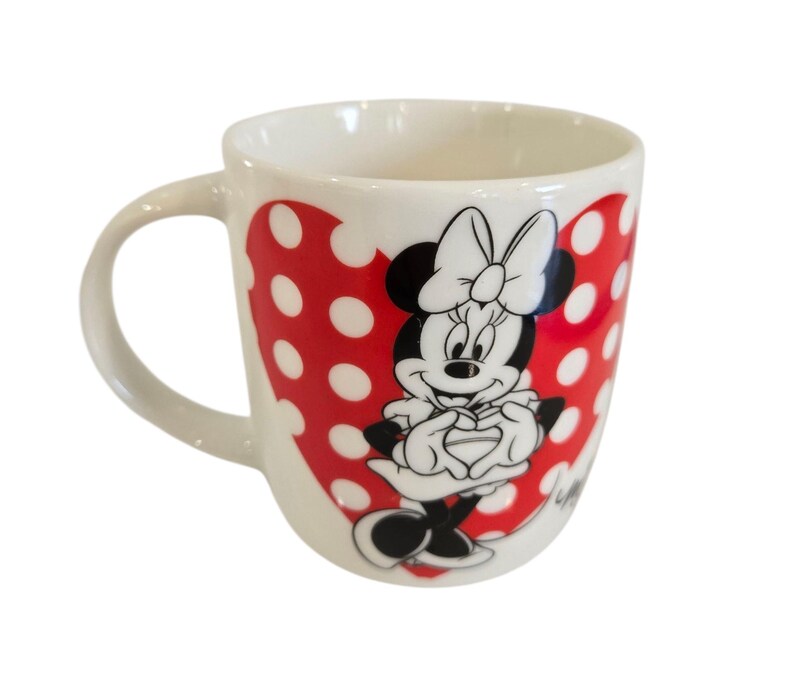 Op de afbeelding: Witte keramische mok met Minnie Mouse. De mok heeft een rood hartontwerp met witte stippen. Minnie Mouse is afgebeeld in zwart, wit en rood, met haar handen voor zich. De mok heeft een gebogen handvat.