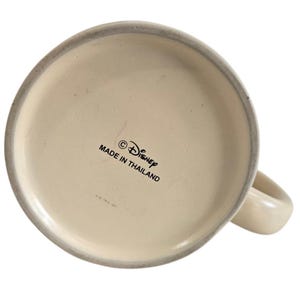 Pode incluir: Caneca de cer&acirc;mica branca com uma al&ccedil;a arredondada. A base da caneca apresenta o s&iacute;mbolo de copyright, a palavra "Disney" e o texto "MADE IN THAILAND" em letras pretas.