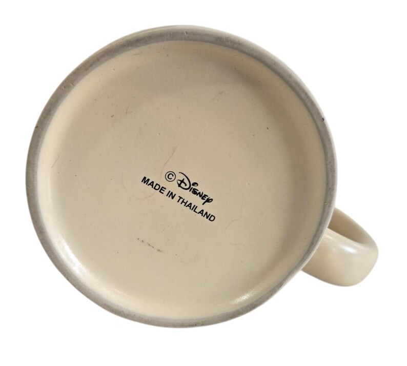 Pode incluir: Caneca de cer&acirc;mica branca com uma al&ccedil;a arredondada. A base da caneca apresenta o s&iacute;mbolo de copyright, a palavra "Disney" e o texto "MADE IN THAILAND" em letras pretas.