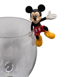 May include: A Mickey Mouse figurine in classic colors of black, red, yellow, and white, perched on the rim of a clear glass. The figure is in a cheerful pose, with one arm raised. The glass has a decorative design near the base.