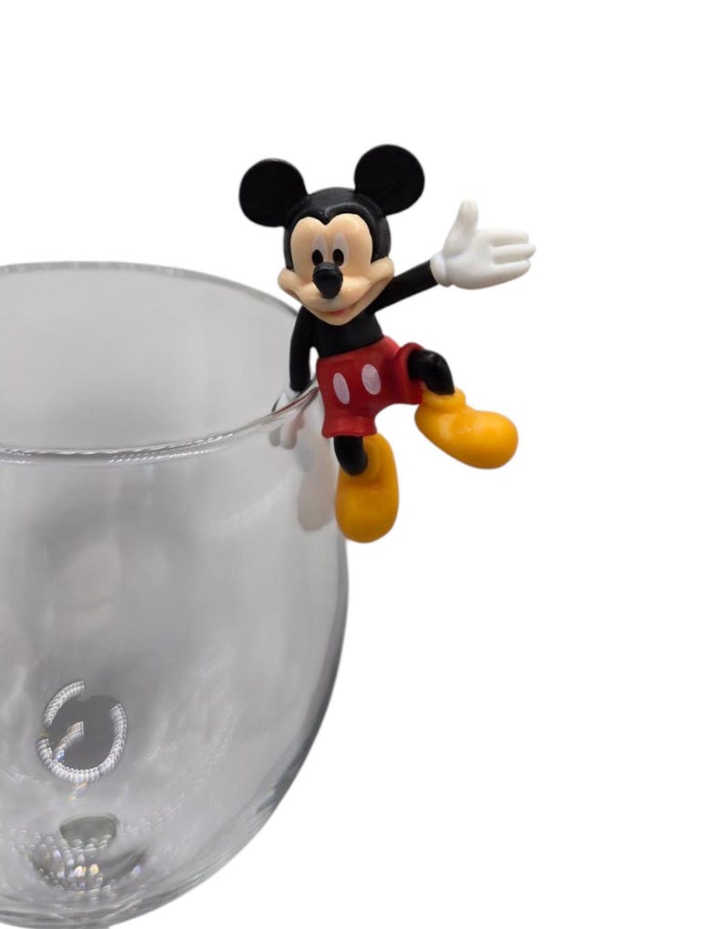 May include: A Mickey Mouse figurine in classic colors of black, red, yellow, and white, perched on the rim of a clear glass. The figure is in a cheerful pose, with one arm raised. The glass has a decorative design near the base.