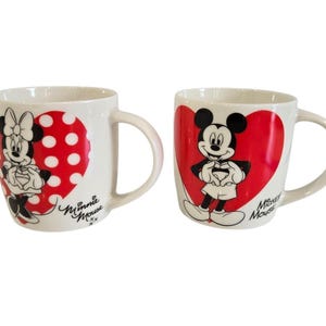 Op de afbeelding: Twee witte keramische mokken met Disney-personages. E&eacute;n mok toont Minnie Mouse in een rode hart met stippen, de andere Mickey Mouse in een rode hart. Beide mokken hebben gebogen handvatten en de woorden "Minnie Mouse" en "Mickey Mouse" in script.