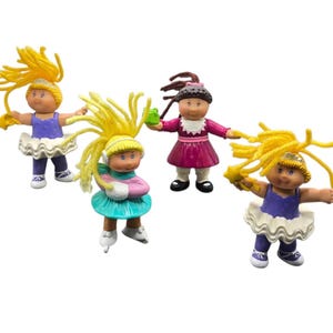 May include: Four Cabbage Patch Kids dolls are shown. Two dolls wear tutus and ballet shoes, one doll wears ice skates, and another doll wears a dress. The dolls have yellow or brown yarn hair and are holding accessories.