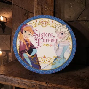 Puede incluir: Un letrero decorativo circular con los personajes animados Anna y Elsa de Frozen. El texto Sisters Forever está impreso en el centro con una fuente estilizada. El borde es azul con patrones florales y detalles en filigrana dorada.