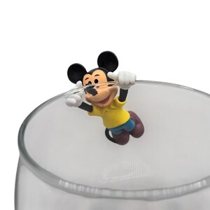 May include: A clear glass with a Mickey Mouse figurine attached to the rim. The Mickey Mouse figure is wearing a yellow shirt, blue shorts, and brown shoes. The figurine is holding onto the rim of the glass with white gloves.