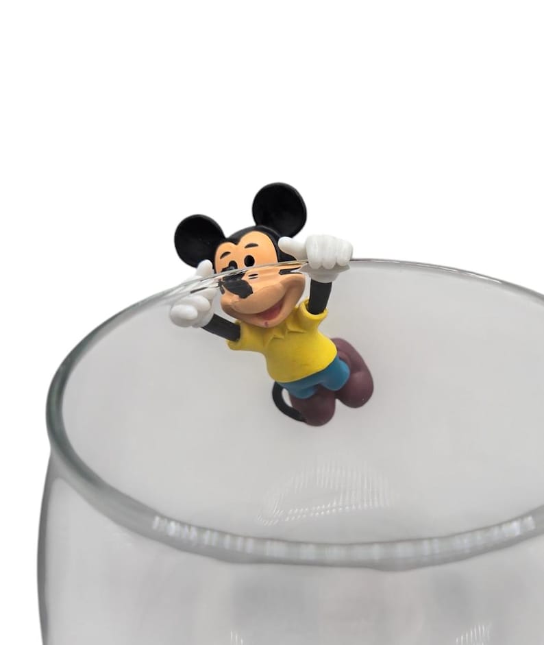 May include: A clear glass with a Mickey Mouse figurine attached to the rim. The Mickey Mouse figure is wearing a yellow shirt, blue shorts, and brown shoes. The figurine is holding onto the rim of the glass with white gloves.