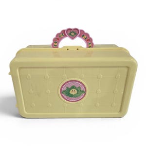 May include: A light yellow plastic carrying case with a pink handle and a circular emblem featuring a cabbage patch kid. The case has a grid pattern with small circles and is likely a vintage toy.