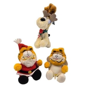 May include: Three plush toys: a reindeer, and two Garfield characters. The reindeer is cream-coloured with brown antlers and a red tongue. One Garfield is dressed as Santa Claus in a red suit. The other Garfield is wearing a white outfit.