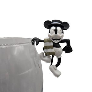 May include: A black and white Mickey Mouse figurine with a black hat and a striped scarf, perched on the edge of a clear glass. The figurine is smiling with its eyes closed. The glass is partially visible.
