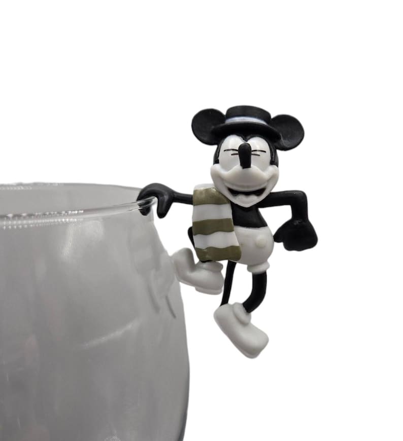 May include: A black and white Mickey Mouse figurine with a black hat and a striped scarf, perched on the edge of a clear glass. The figurine is smiling with its eyes closed. The glass is partially visible.