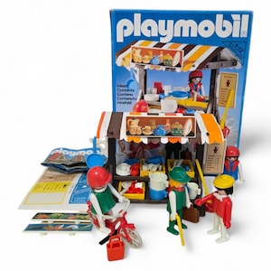 Puede incluir: Juego de juguetes Playmobil con un puesto de mercado con toldo a rayas, figuras en miniatura y accesorios. El juego incluye una bicicleta roja, figuras con sombreros y una caja con el logotipo de Playmobil. La caja tiene texto en varios idiomas.