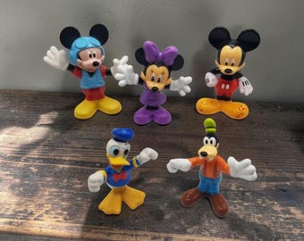 May include: A set of five plastic character figurines including Mickey Mouse wearing a blue aviator cap, Minnie Mouse in a purple dress with a bow, a standard Mickey Mouse, Donald Duck in a blue sailor shirt, and Goofy in an orange shirt and blue pants.
