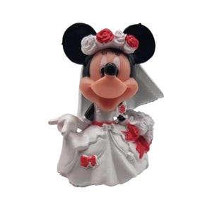 May include: A figurine of Minnie Mouse in a white wedding dress with red accents, a veil, and a floral headpiece. The figure has black ears and a smiling expression, holding a bouquet.