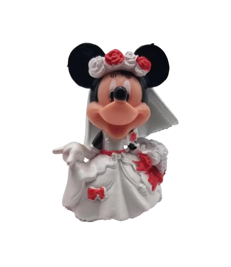 May include: A figurine of Minnie Mouse in a white wedding dress with red accents, a veil, and a floral headpiece. The figure has black ears and a smiling expression, holding a bouquet.