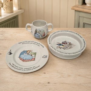 Peut inclure: Un ensemble de vaisselle pour enfant en céramique blanche composé de trois pièces illustrées de Peter Rabbit avec du texte. Le set comprend un mug à deux anses, une assiette plate et un bol creux, ornés de scènes colorées et de texte imprimé.