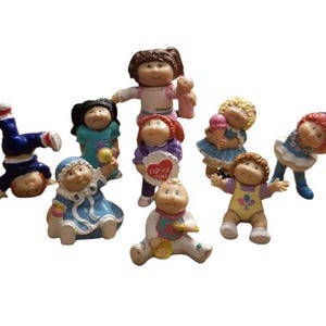 May include: A collection of vintage Cabbage Patch Kids figurines in various poses. The dolls are dressed in colorful outfits and have different hairstyles. One doll is doing a handstand, while another holds a heart-shaped sign that says "I Love You."