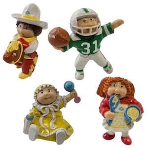 Op de afbeelding: Vier Cabbage Patch Kids beeldjes. Eén draagt een sombrero en houdt een paard vast, een ander is een American footballspeler met nummer 31, een derde houdt een rammelaar vast en de laatste houdt een lepel en een kom vast.