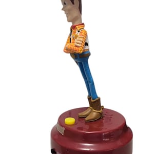 May include: A Woody action figure from the Toy Story franchise. The figure is wearing a cowboy hat, plaid shirt, blue jeans, and brown boots. The figure is attached to a red base with a yellow button.